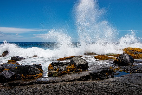 a wave crashes high into the air against lava rocks