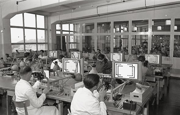 people sit working at desks in a room with large windows