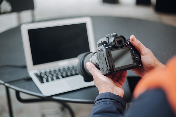 hands hold a camera in front of a laptop set on a round table