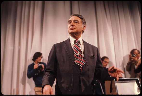 a man in a suit-stands in front of three people with cameras and a curtain