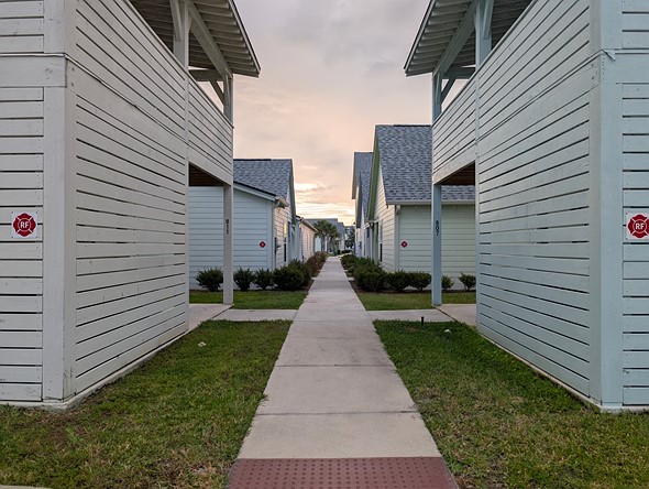 a sidewalk stretches between two rows of pastel colored houses with a faint glow in the cloudy sky