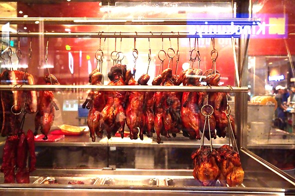 a line of whole cooked ducks hang from a rack behind a glass window in a market
