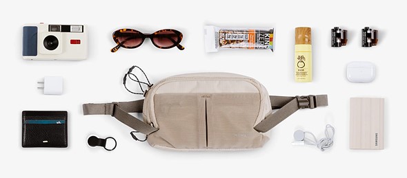 a camera snacks wallet and other items are laid out on a white table around a tan sling