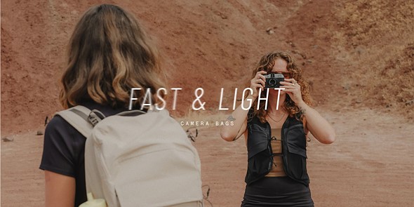 a person stands holding a camera up to her face while another faces away with a white backpack on
