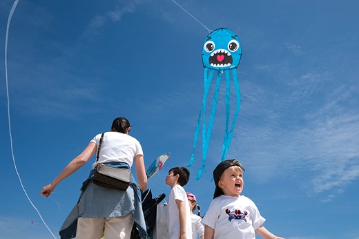 a blue kite with a silly face flies over a group and child making a similar face