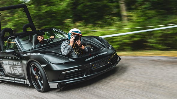 a person with a camera held to their face sits in the front trunk of a black porsche while it speeds down a track