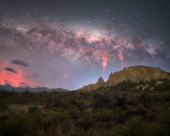 the milky way and red sprites glow above a rock formation and mountains