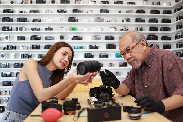 a woman holds a lens up while a man wearing black gloves looks at it in a camera shop