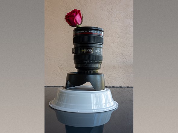 a mug that looks like a lens with a red rose in it is resting on a lens hood and upside down gray bowl
