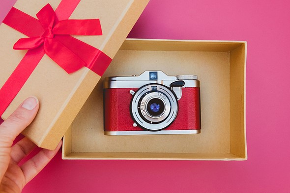 a red and silver camera is in a brown box while a hand holds the lid with a red bow
