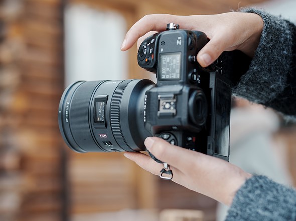 hands hold a black nikon camera with viltrox lens in front of a wooden building