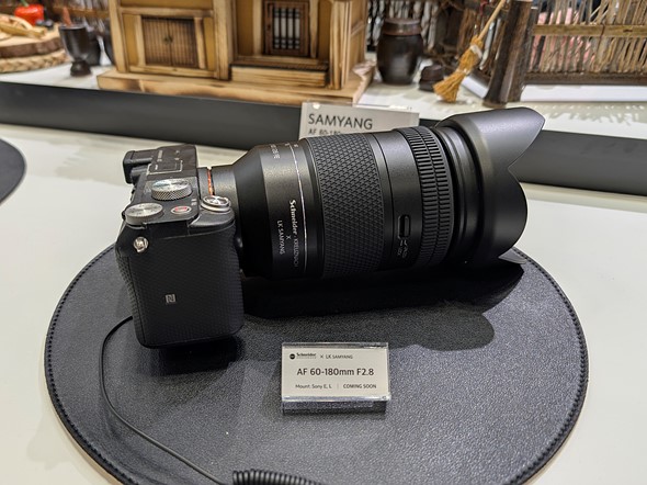 a black lens on a camer sits on a round mat on a white table with model houses behind it