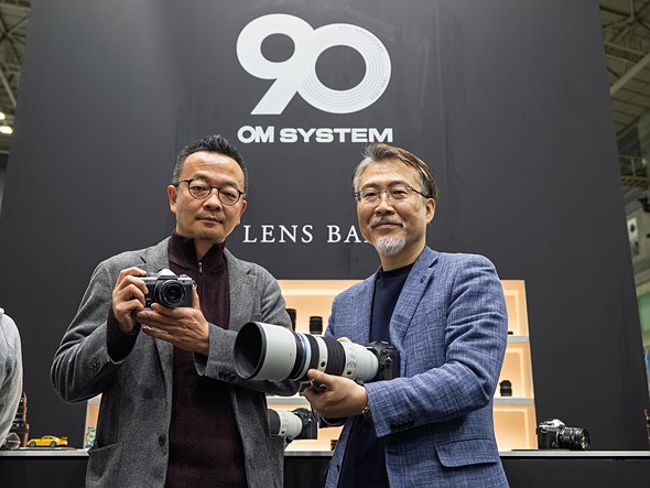 two men in suits holding cameras stand in front of a black wall with white text