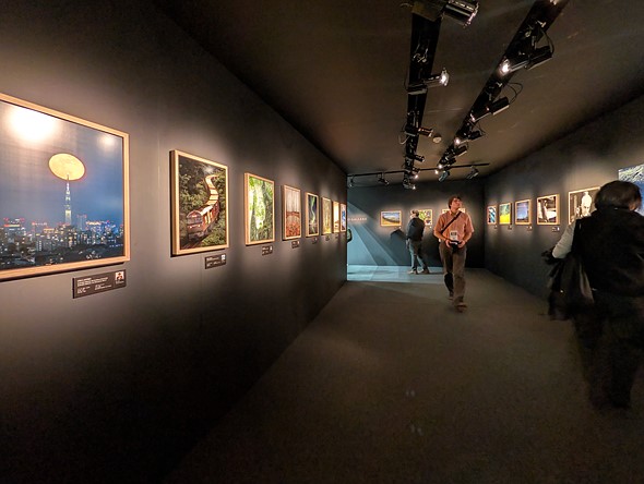 framed photographs hang on the walls of a black gallery space with people walking around