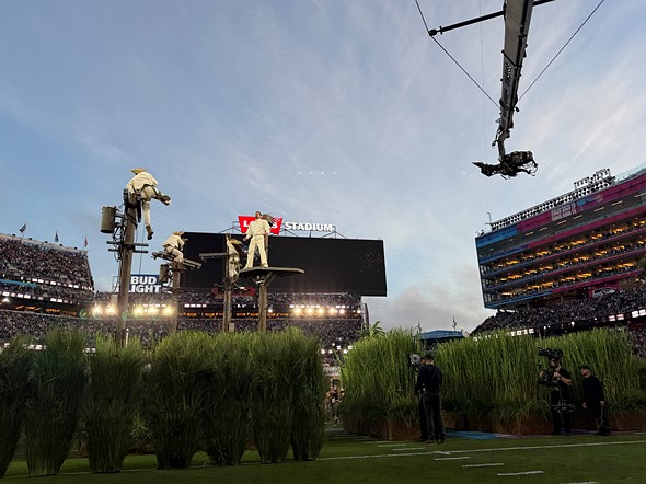 people dressed in white stand on poles amidst tall fake grass placed in a big football stadium