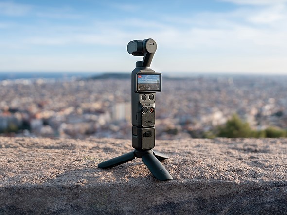 a gimbal camera with mini tripod at the bas stands on a rocky ledge with a blurred out city in the distance