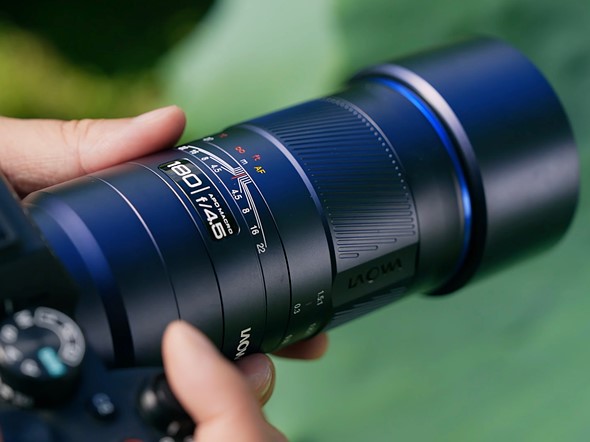 fingers hold a black laowa telephoto macro lens over a blurred green background