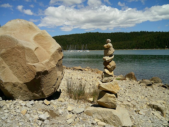 Stack of balanced rocks on a rocky lakeshore near a large boulder with calm water forested hills and sailboats in the background