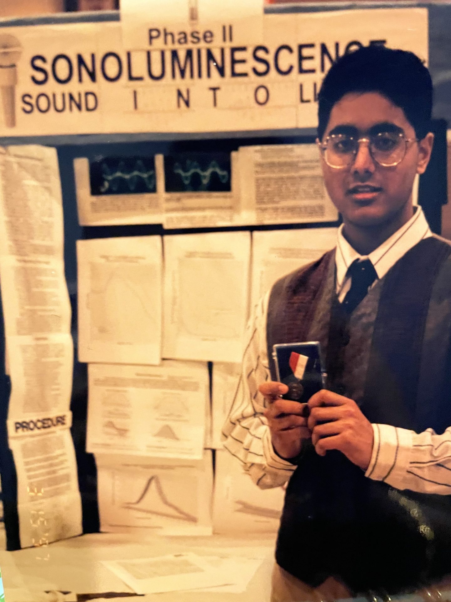 Person wearing glasses and a vest stands in front of a science fair project display board about sonoluminescence, holding an award. The board features charts, graphs, and printed documents.