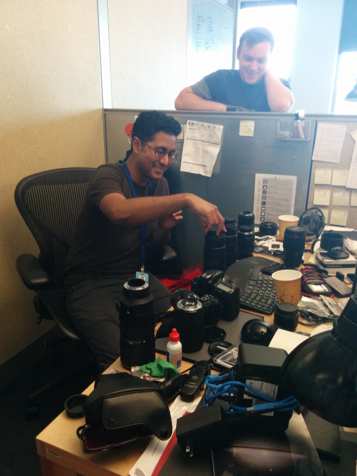 Person seated at a cluttered desk in an office cubicle, handling camera equipment alongside various lenses, coffee cups, and office supplies; another person stands nearby.