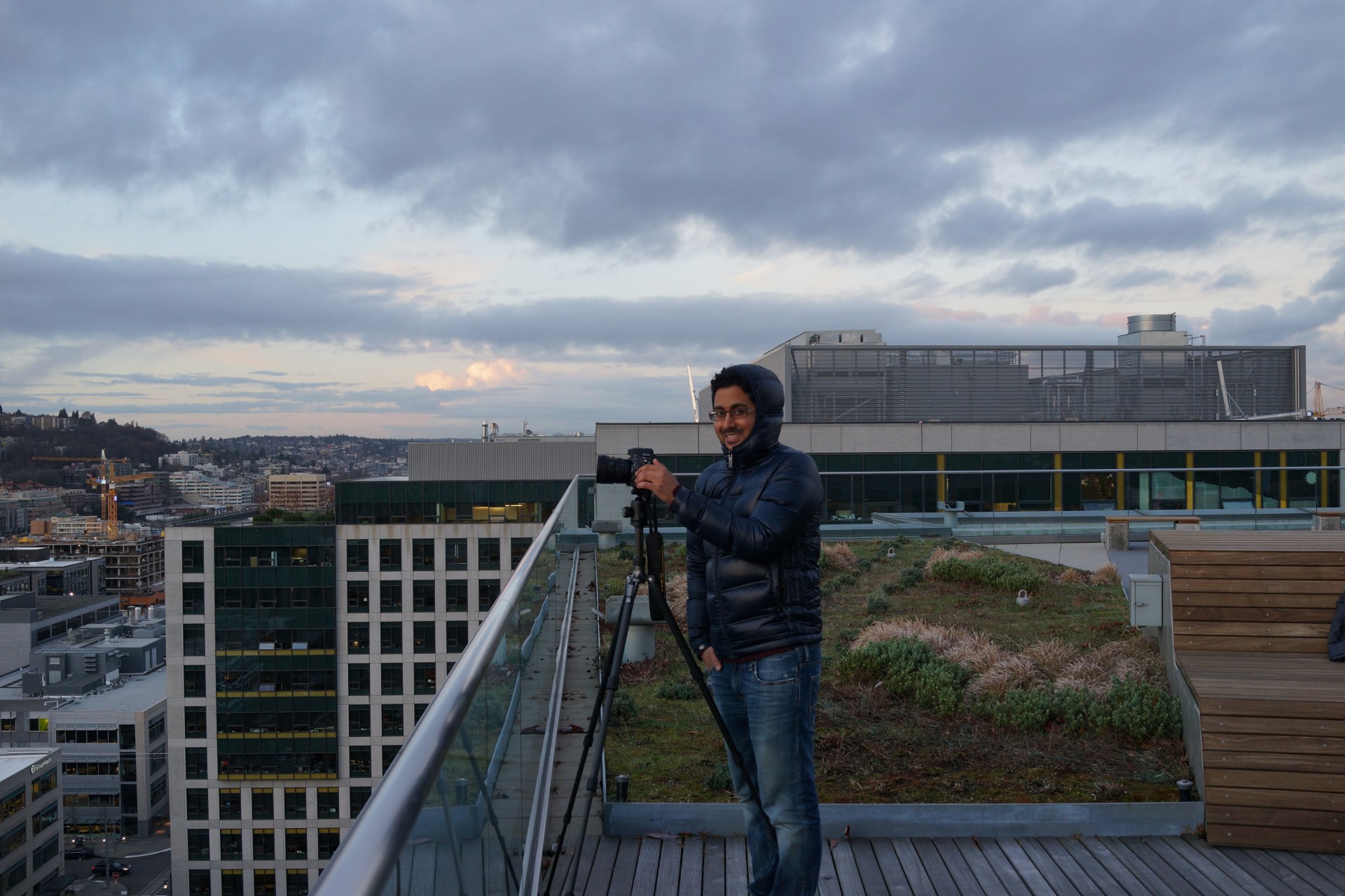 Person wearing a hooded jacket stands on a rooftop deck, holding a camera mounted on a tripod; city buildings and a cloudy sky are in the background.