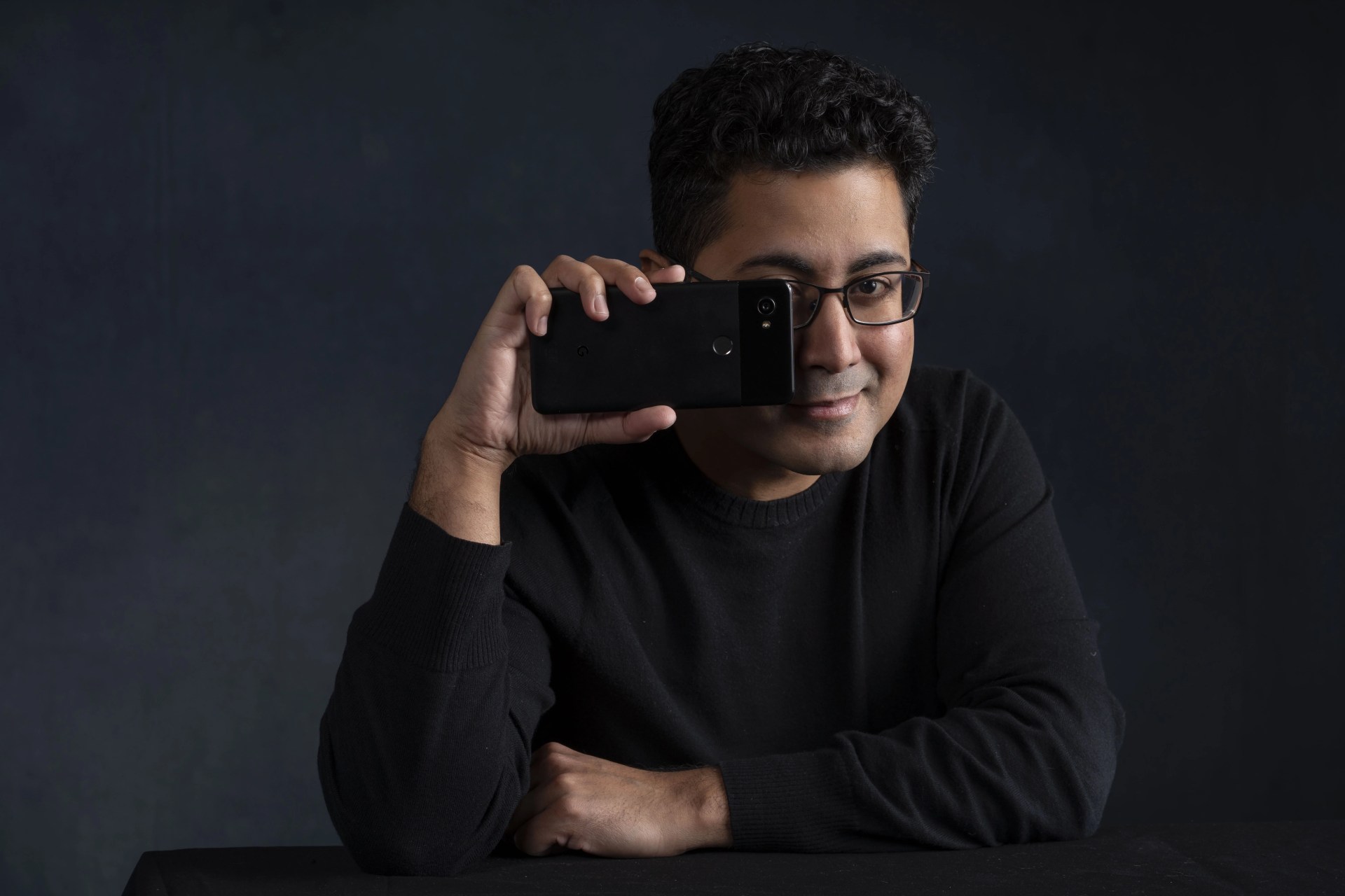 Person wearing glasses and a black sweater smiles while holding a smartphone in front of their face, covering one eye, with a dark background.